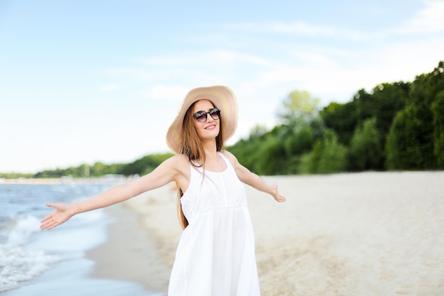모자, 선글라스, 손을 벌린 채 바다 해변에 서서 자유로운 행복을 누리며 웃고 있는 행복한 여성. 여행 중 자연을 즐기는 흰색 여름 드레스를 입은 다문화 여성 모델의 초상화