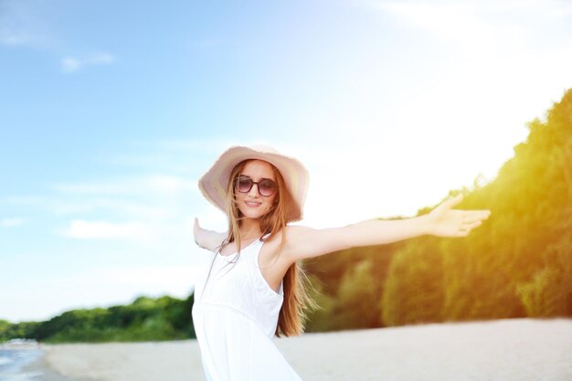 Photo happy smiling woman in free happiness bliss on ocean beach standing with a hat sunglasses and open hands portrait of a multicultural female model in white summer dress enjoying nature