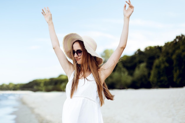 Photo happy smiling woman in free happiness bliss on ocean beach standing with a hat, sunglasses, and rasing hands. portrait of a multicultural female model in white summer dress enjoying nature during trav