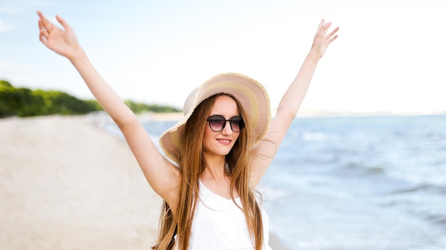 Happy smiling woman in free happiness bliss on ocean beach standing with a hat, sunglasses, and rasing hands. Portrait of a multicultural female model in white summer dress enjoying nature during trav