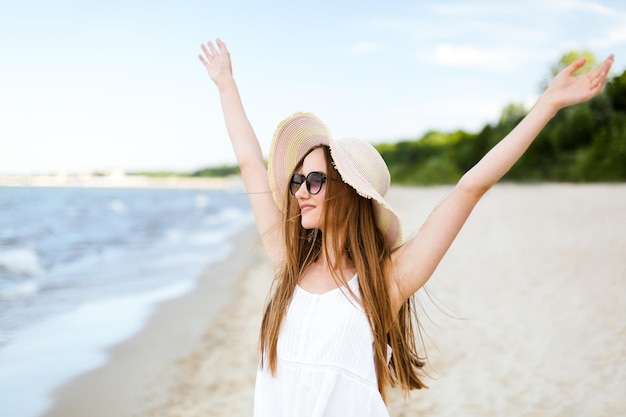 Счастливая улыбающаяся женщина в свободном блаженстве счастья на океанском пляже, стоящая в шляпе, солнцезащитных очках и поднятых руках. Портрет мультикультурной женщины-модели в белом летнем платье, наслаждающейся природой во время трава