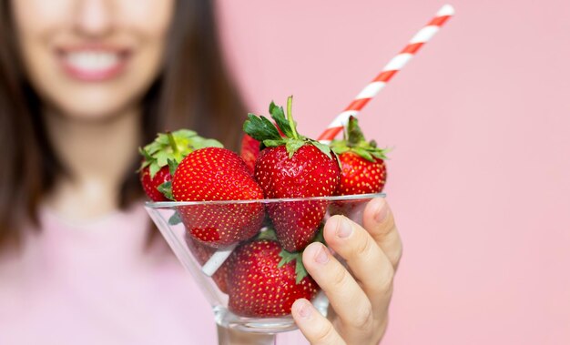 happy smiling woman holding glass bowl with fresh strawberries in hand and drinking straw in mouth