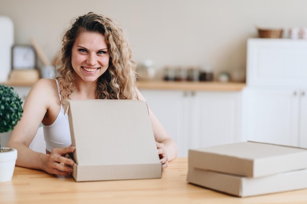 Happy smiling woman opens a delivery box A woman sits at a wooden table in a bright home interior