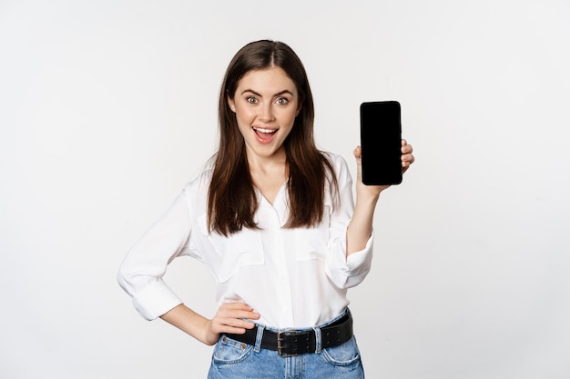 Foto donna sorridente felice che mostra l'interfaccia dell'app per smartphone, lo schermo del telefono cellulare, dimostrando l'offerta promozionale, in piedi su sfondo bianco.