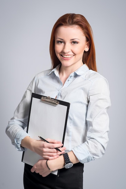 Happy smiling woman in strict clothes and glasses holding a clipboard on grey