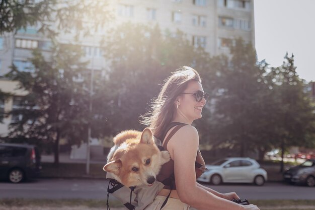 행복한 미소 짓는 여성 여행자가 특별한 배낭에 Welsh Corgi Pembroke 강아지와 함께 도시 공원에서 전기 스쿠터를 타고 있습니다