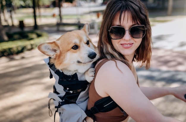 Happy smiling woman traveler is riding her electro scooter in city parkland with dog Welsh Corgi Pembroke in a special backpack