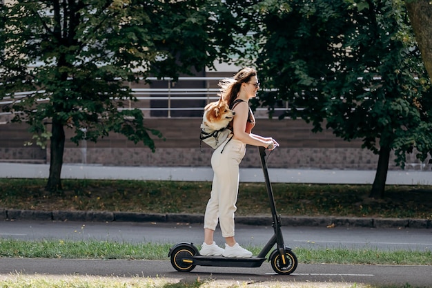 Happy smiling woman traveler is riding her electro scooter in\
city parkland with dog welsh corgi pembroke in a special\
backpack