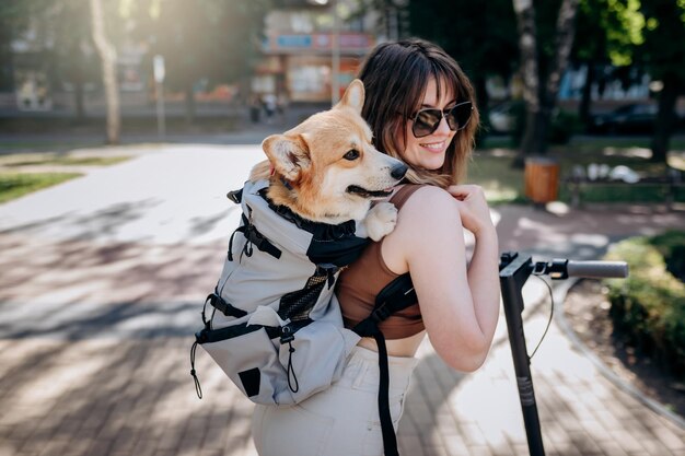 행복한 미소 짓는 여성 여행자가 특별한 배낭에 Welsh Corgi Pembroke 강아지와 함께 도시 공원에서 전기 스쿠터를 타고 있습니다