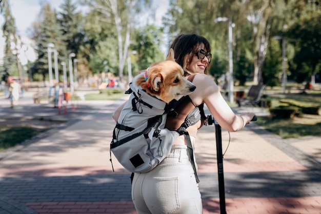 Happy smiling woman traveler is riding her electro scooter in city parkland with dog Welsh Corgi Pembroke in a special backpack