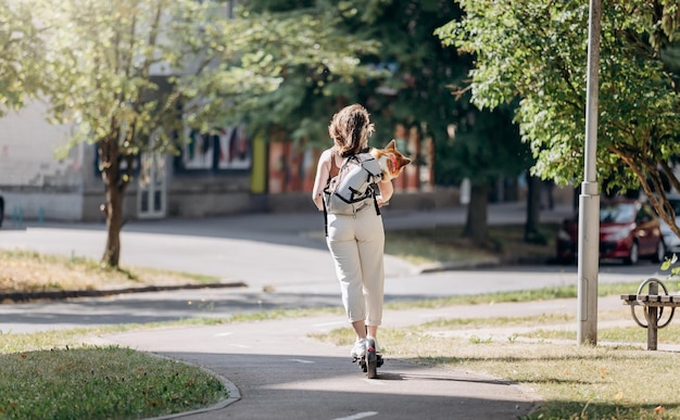 Happy smiling woman traveler is riding her electro scooter in city parkland with dog Welsh Corgi Pembroke in a special backpack