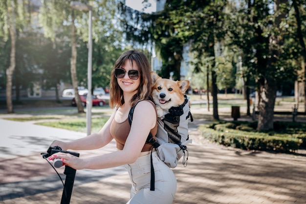 행복한 미소 짓는 여성 여행자가 특별한 배낭에 Welsh Corgi Pembroke 강아지와 함께 도시 공원에서 전기 스쿠터를 타고 있습니다