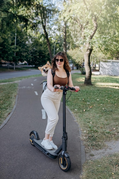 Happy smiling woman traveler is riding her electro scooter in city parkland with dog Welsh Corgi Pembroke in a special backpack