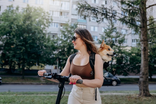행복한 미소 짓는 여성 여행자가 특별한 배낭에 Welsh Corgi Pembroke 강아지와 함께 도시 공원에서 전기 스쿠터를 타고 있습니다
