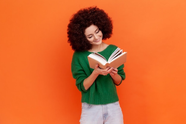 Happy smiling woman with Afro hairstyle wearing green casual style sweater reading book with interesting plot, studying with pleasure. Indoor studio shot isolated on orange background.