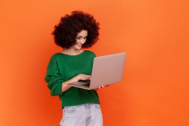 Happy smiling woman with afro hairstyle wearing green casual
style sweater working on portable computer looking at display with
positive expression indoor studio shot isolated on orange
background