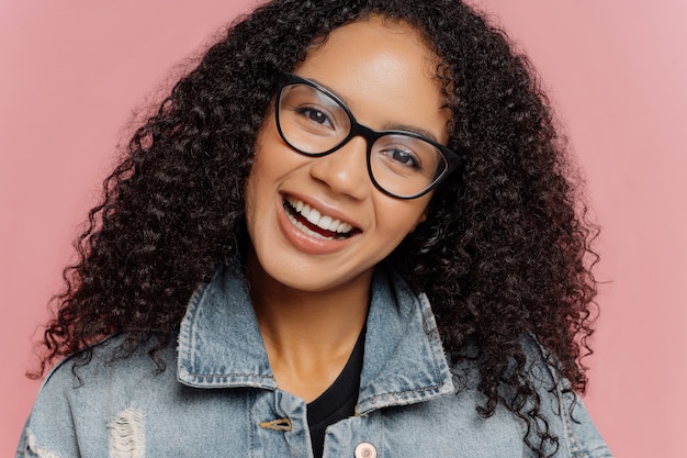 Happy smiling woman with dark curly Afro hairstyle, tilts head, wears optical glasses and denim jacket