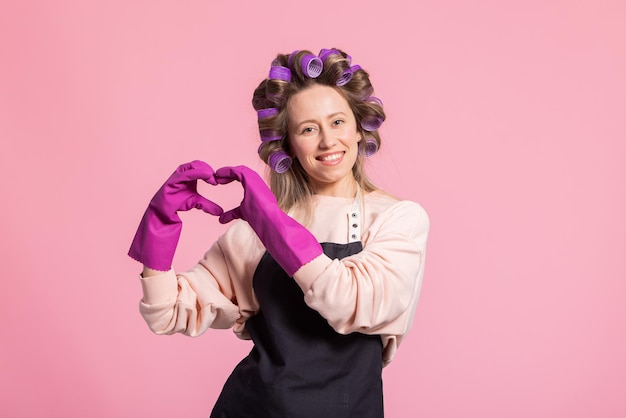 Happy smiling woman with rollers on her head wearing rubber protective gloves shows heart to camera beautiful white smile pink studio background