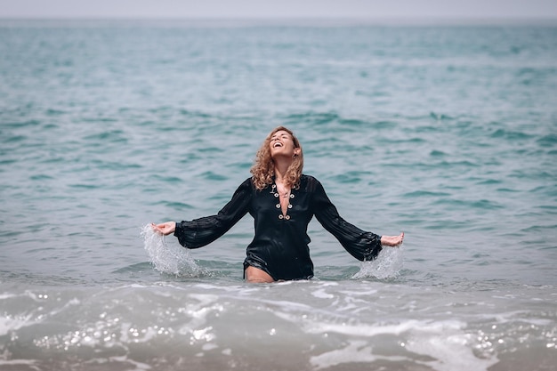 Happy smiling woman with thick blond wavy hair, wearing black long shirt stands in the sea near the shore playing with water, splashes and splatter.