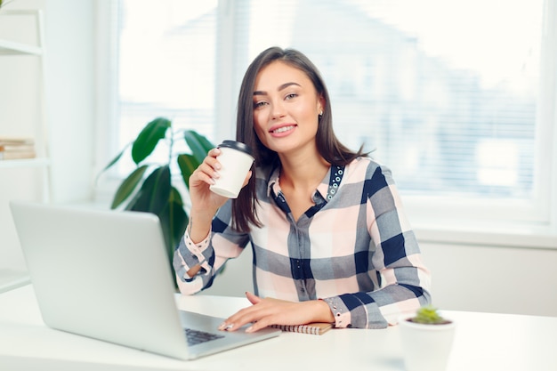 幸せな笑顔の女性のラップトップで働くとコーヒーを飲む