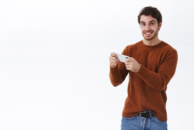 Счастливый улыбающийся молодой 25-летний мужчина с бородой использует смартфон для записи видео, смотрит прямую трансляцию онлайн, держа мобильный телефон горизонтально, играя в игру и взволнованно глядя на камеру на белом фоне