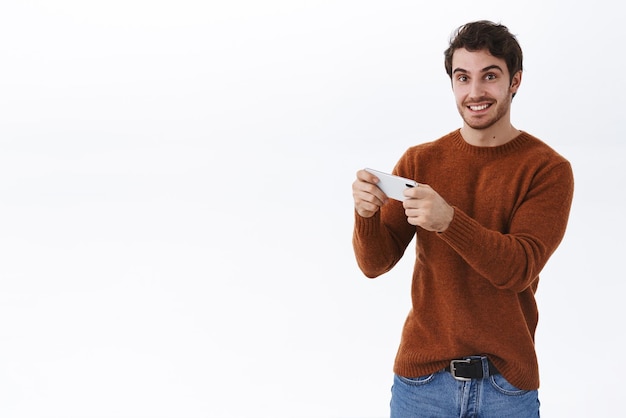 Felice sorridente giovane 25s uomo con la barba che utilizza lo smartphone per registrare video guardando lo streaming live online tenendo il telefono cellulare orizzontalmente giocando e guardando eccitato sfondo bianco della fotocamera