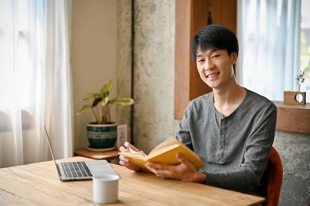 행복하고 웃는 젊은 아시아 남자가 책과 노트북을 들고 커피숍에 앉아 있다