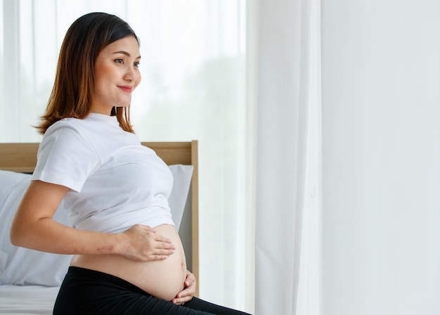 Happy smiling young beautiful Asian pregnant woman sitting on a bed looking and touching her belly with care and love. Expecting for a strong and healthy baby.