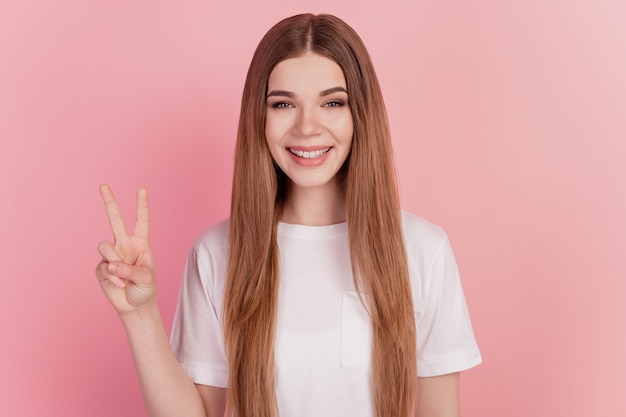 Фото Счастливая улыбающаяся молодая милая женщина, показывающая жест победы двумя пальцами на розовой стене