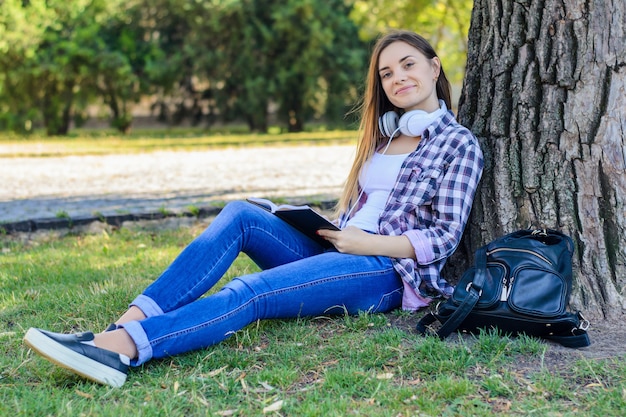 Счастливая улыбающаяся молодая девушка в повседневной одежде с наушниками на шее сидит на траве и читает книгу