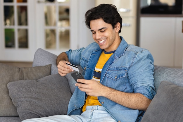 Happy smiling young male or student with credit card uses mobile phone making profitable purchases in the Internet sitting on the couch at home