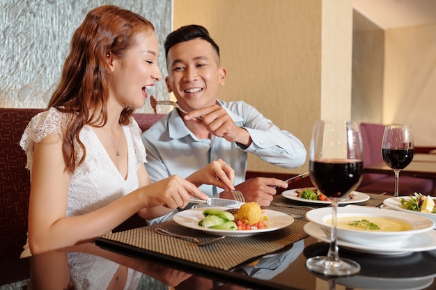 Happy smiling young man feeding girlfriend with piece of meat from his plate