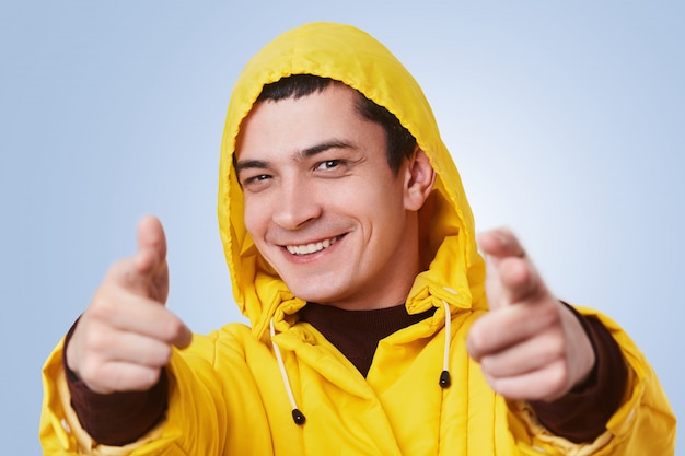 Happy smiling young man wears yellow anorak and hood, points at you with glad expression, chooses someone, isolated over blue