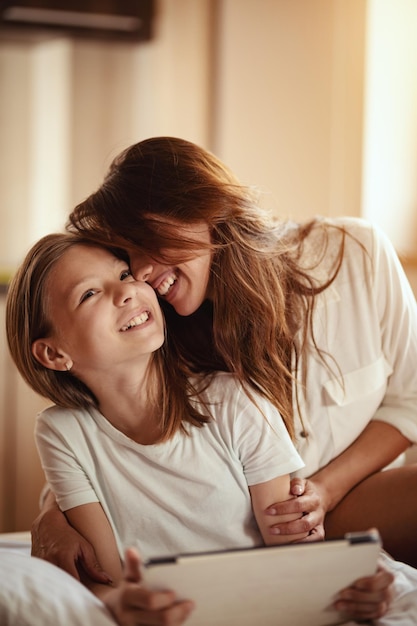 Happy smiling young mother is embracing her daughter, who is holding a digital tablet, while they are sitting on bed at home.