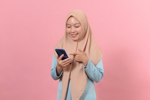 Happy smiling young muslim woman hold smart phone in hands, looking at display screen texting messages using online social media apps standing isolated on pink background