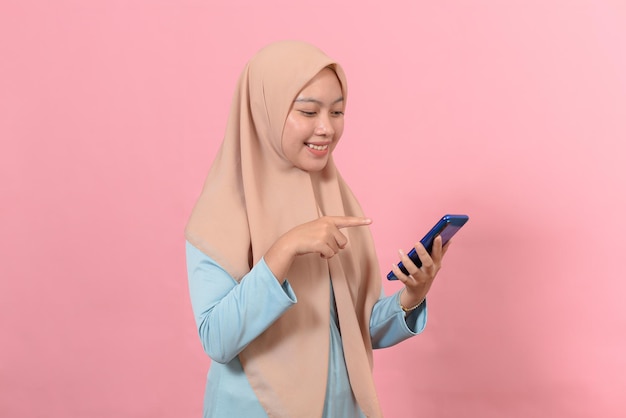 Happy smiling young muslim woman hold smart phone in hands, looking at display screen texting messages using online social media apps standing isolated on pink background