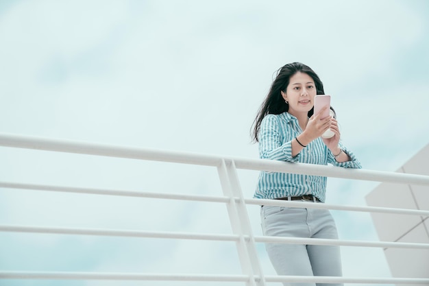 happy smiling young office lady having coffee break time and stay alone on rooftop of office building skyscraper. joyful business woman worker texting chatting online with friend leaning on railing