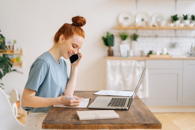 Happy smiling young redhead woman is talking with client by mobile phone making notes in documents