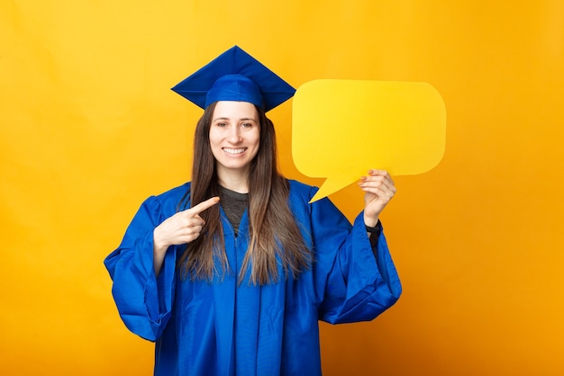 Happy smiling young woman in blue robe pointing at empty yellow speech bubble