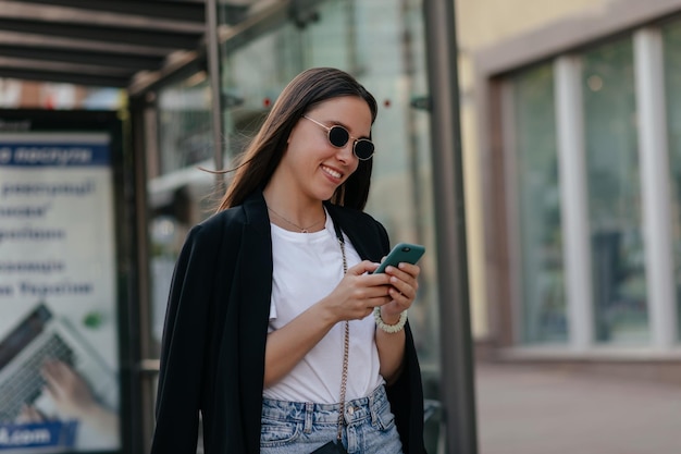 暗い眼鏡をかけた幸せな笑顔の若い女性は、暖かい春の日にスマートフォンをスクロールして街を歩いています素敵な女性は屋外でスマートフォンを使用しています