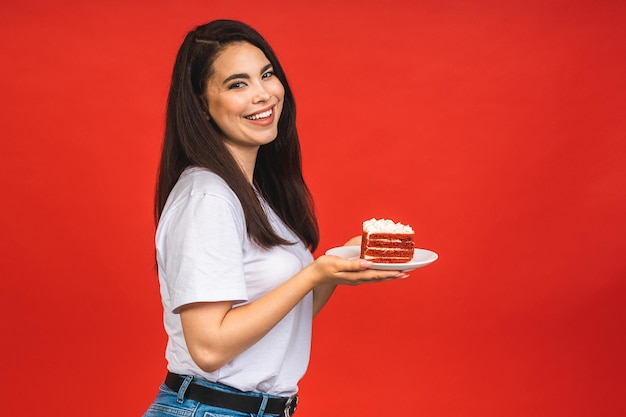 Happy smiling young woman eating the cake isolated over red background Brunette lady holding a birthday cake