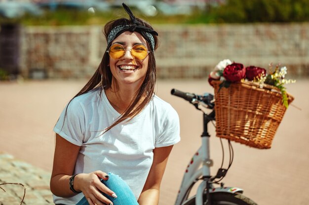 幸せな笑顔の若い女性は、フラワーバスケット付きの自転車の横で、夏の晴れた日に楽しんでいます。