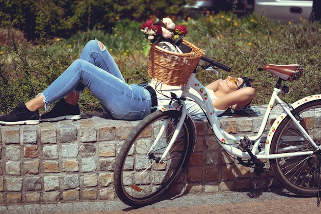 Happy smiling young woman is enjoying in a summer sunny day reposing on little wall in a city, beside the bike with flower basket.