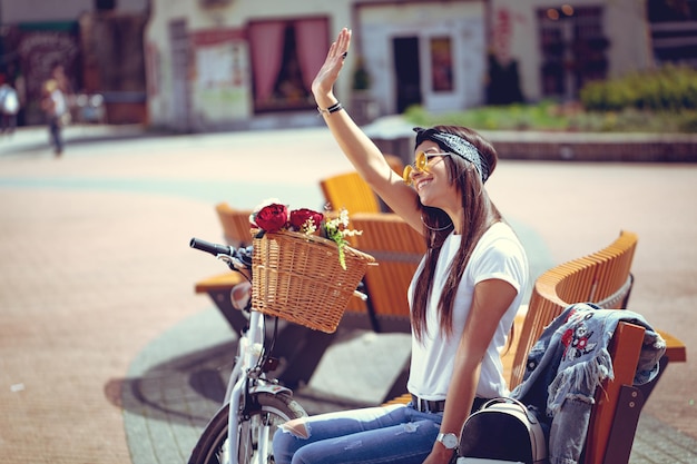 Happy smiling young woman is enjoying in a summer sunny day, sitting on a city bench, beside the bike with flower basket and wave to somebody.