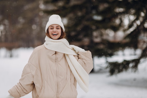 幸せな笑顔の若い女性の肖像画は、コート スカーフ 帽子とミトンを着て、雪の降る冬の公園で冬の天気を楽しんでいます