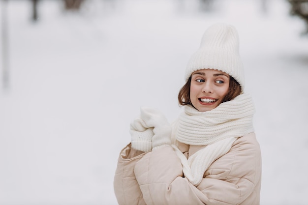 Portrait di giovane donna sorridente felice vestita con cappotto, sciarpa, cappello e guanti, gode del tempo invernale nel parco invernale innevato