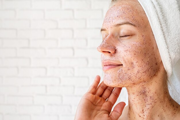 Happy smiling young woman in white bath towels applying scrub on her face