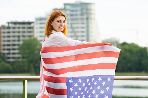 Happy smiling young womanl with USA national flag on her shoulders with high city buildings in background celebrating United States independence day