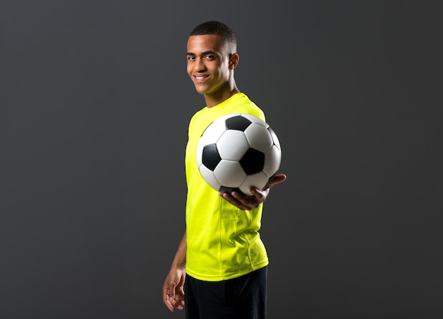 Happy soccer player man with dark skinned playing catching a ball with his hands on dark background