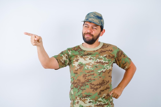 Happy soldier is pointing left with forefinger and putting other hand on waist on white background